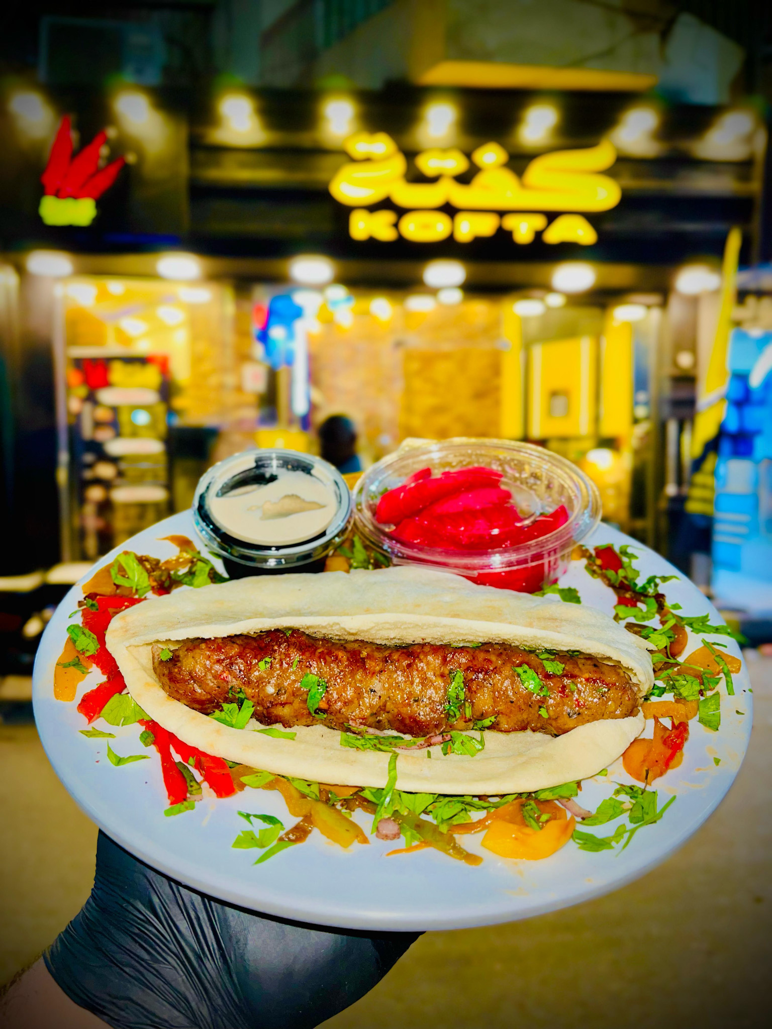 A freshly grilled kofta sandwich served on a plate with pickled vegetables and tahini sauce, in front of a bright yellow-lit Kofta restaurant in Luxor, Egypt.