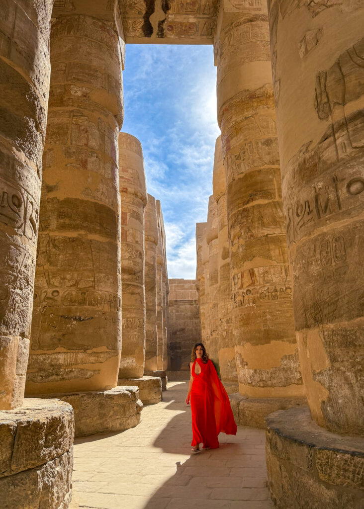 A woman in a flowing red dress walks through the massive sandstone columns of Karnak Temple in Luxor, Egypt, with intricate hieroglyphs and carvings surrounding her.