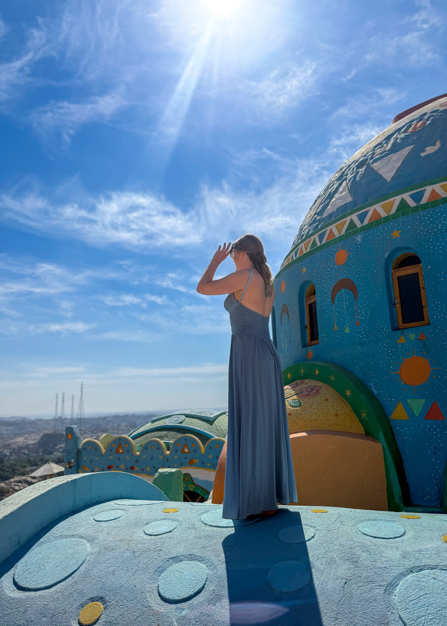 A woman in a flowing blue dress walking on the vibrant, dome-shaped rooftops of Kato Dool in Aswan, surrounded by the bright colors of Nubian architecture under a clear blue sky.