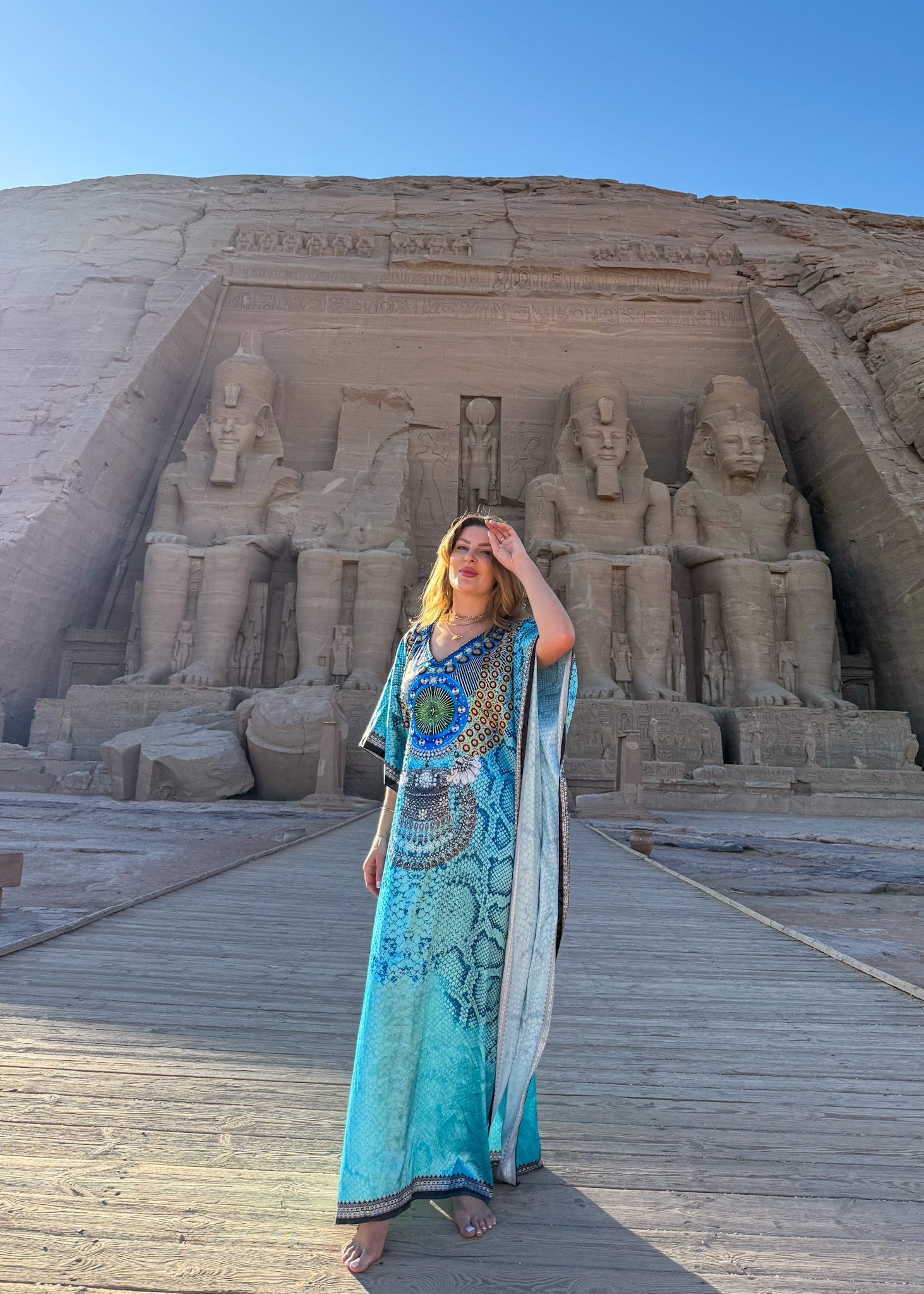 A woman in a vibrant blue kaftan standing in front of the grand statues of Abu Simbel, bathed in the morning sun.