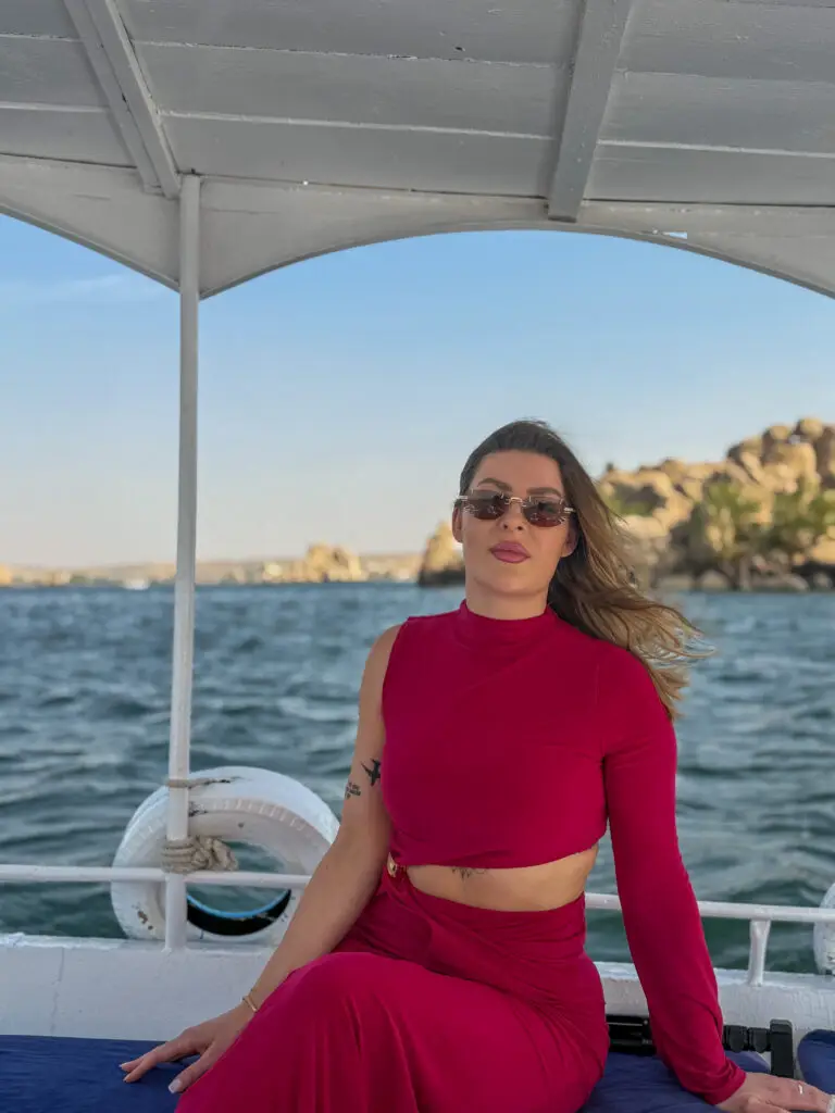 A woman in a red dress enjoying a boat ride on the Nile River in Aswan, Egypt, with scenic views of golden rocks and blue waters in the background.