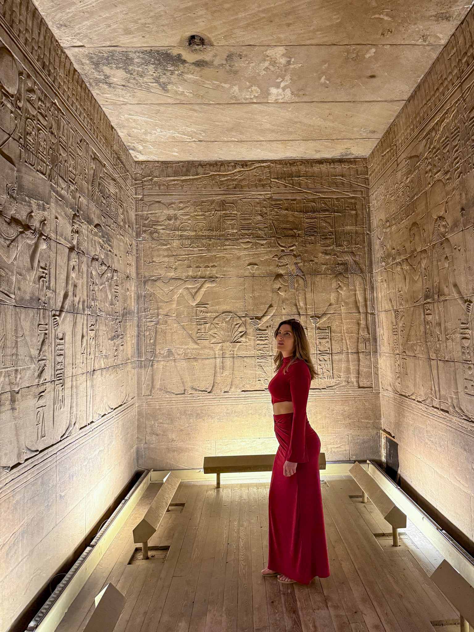 A woman in a striking red dress standing inside an ancient temple in Aswan, Egypt, surrounded by intricate hieroglyphics and carvings.