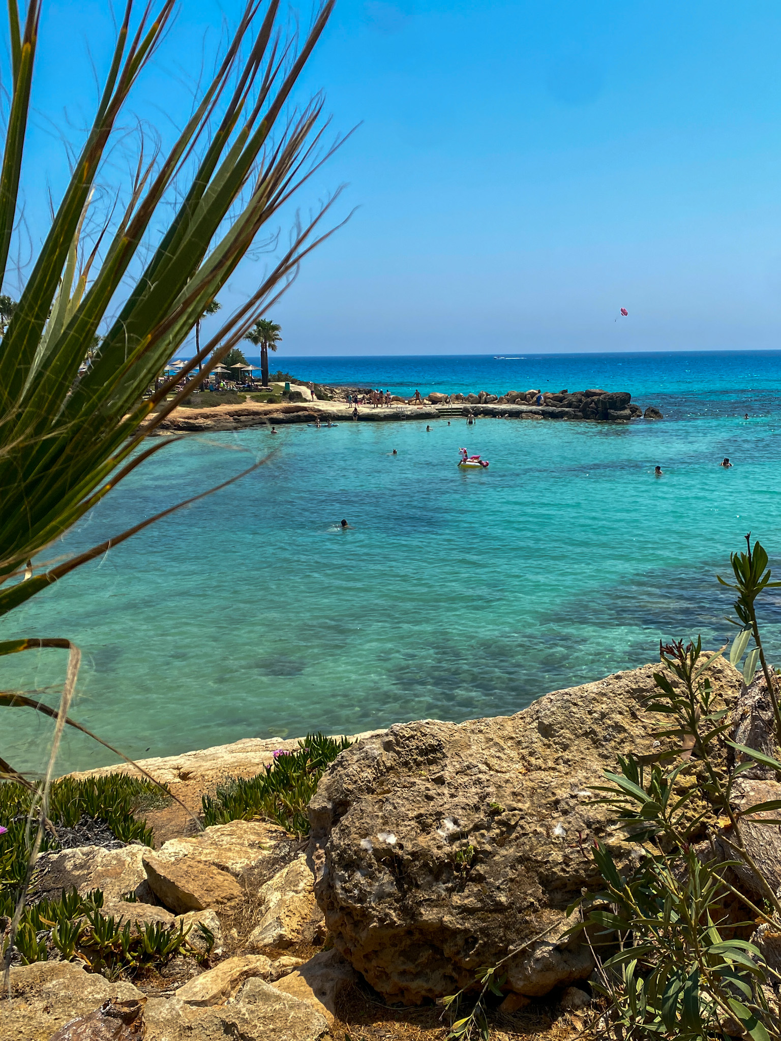 A view of Nissi Beach with crystal-clear turquoise waters, golden sand, and lush greenery under a bright summer sky.