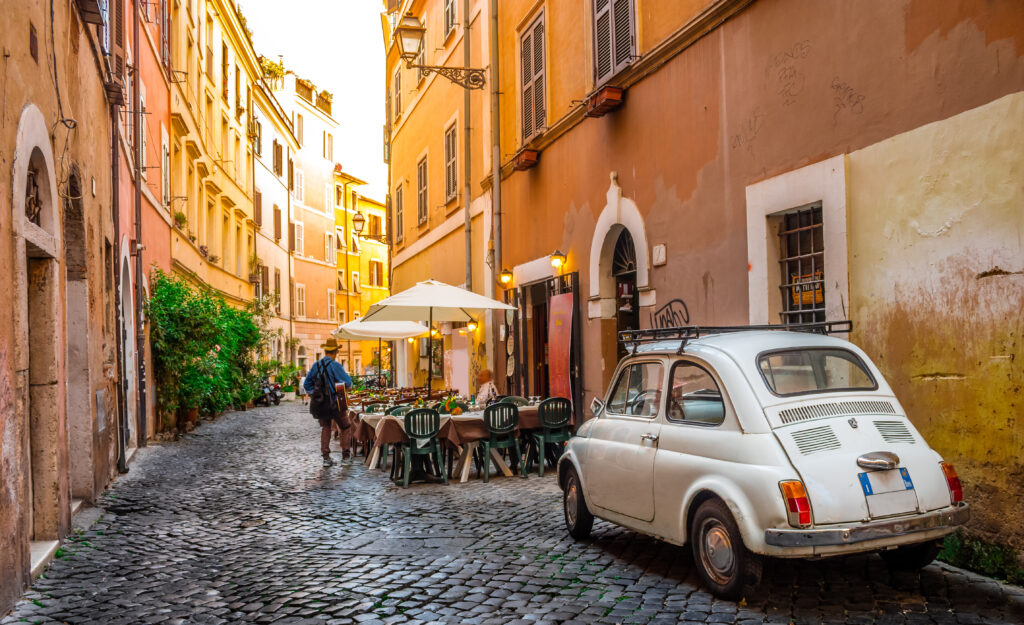 A charming cobblestone street in Trastevere, Rome, featuring a classic Fiat 500, warm-colored buildings, and a cozy outdoor café setting.