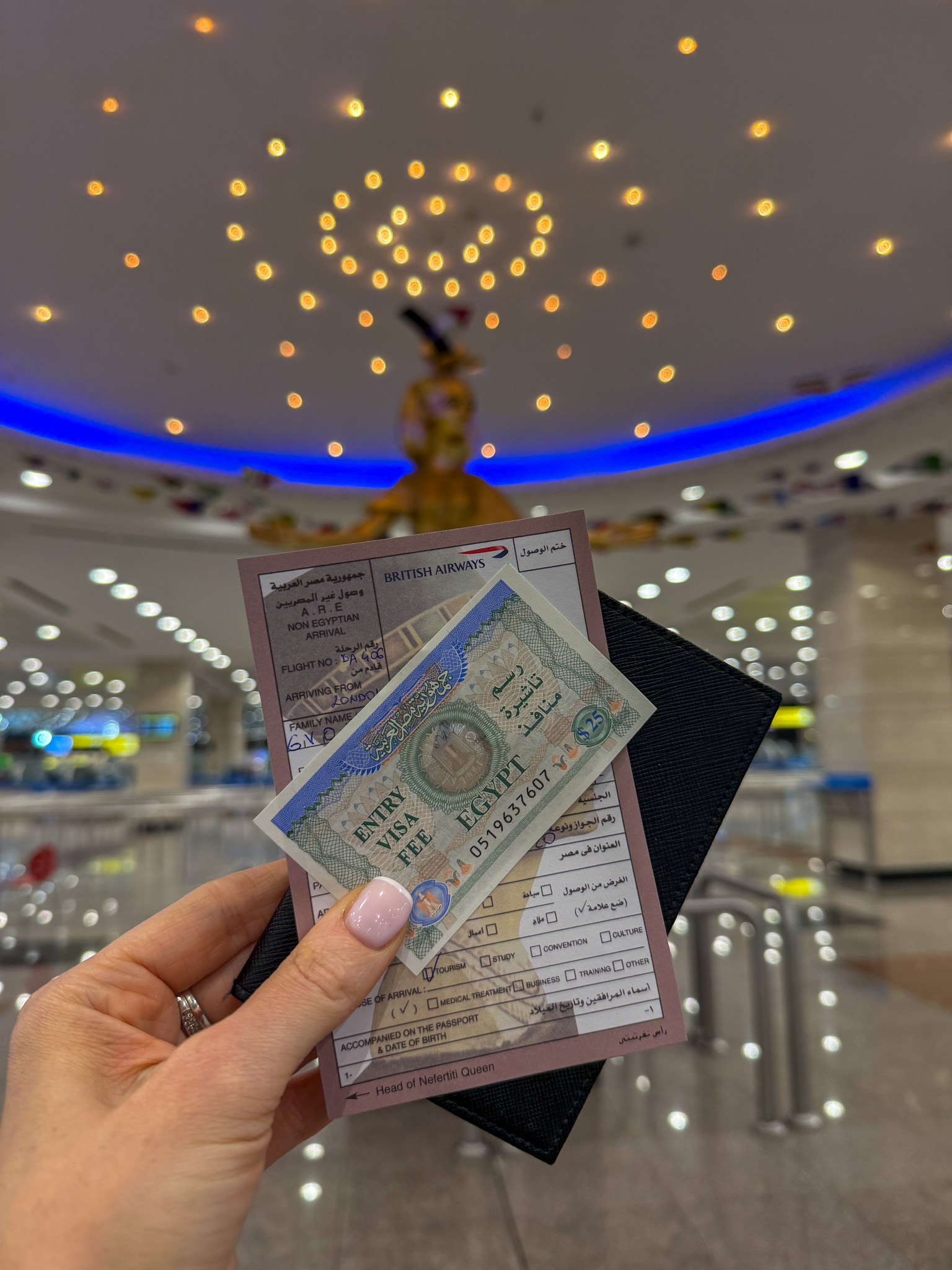 A close-up of a hand holding an Egypt visa on arrival, a passport, and an entry form at an airport with golden lighting and a Pharaoh statue in the background.