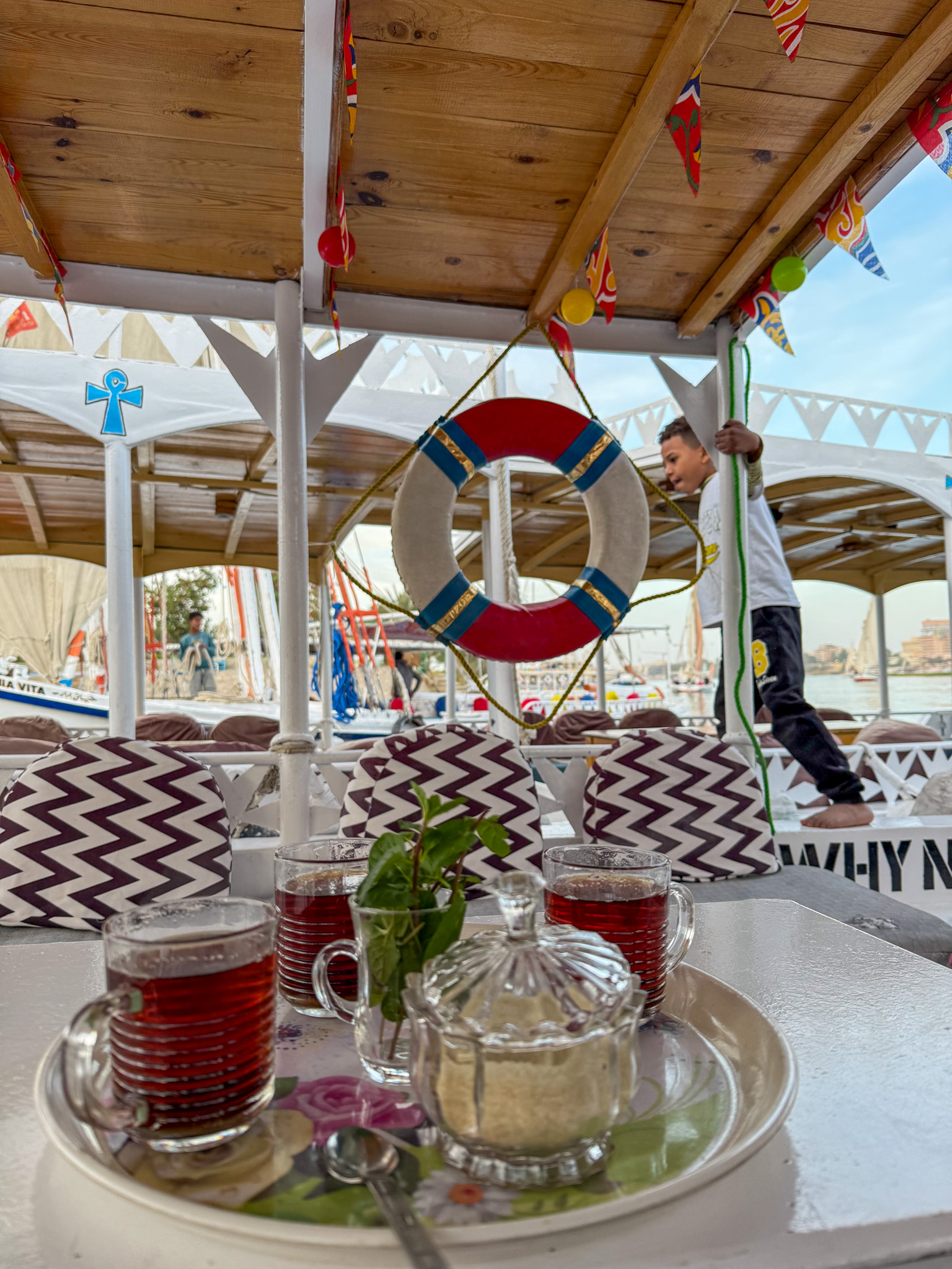 A tray with Egyptian tea, fresh mint, and sugar served on a traditional boat ride along the Nile River, with a vibrant deck and a local boatman in the background.