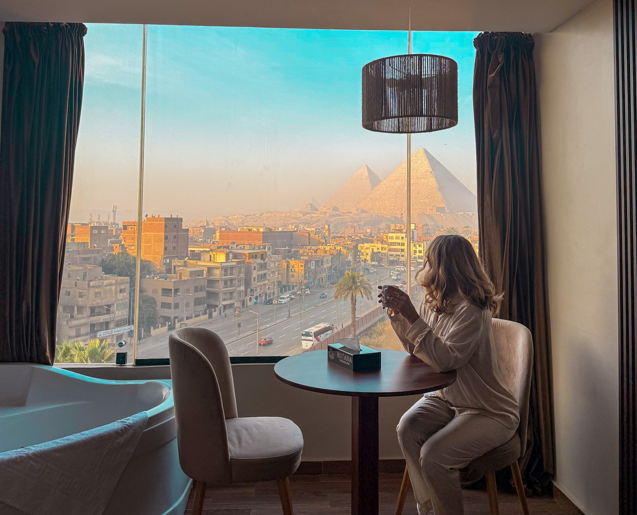 A woman enjoying a cup of coffee in a hotel room with a breathtaking panoramic view of the Giza Pyramids through a large window at sunrise.