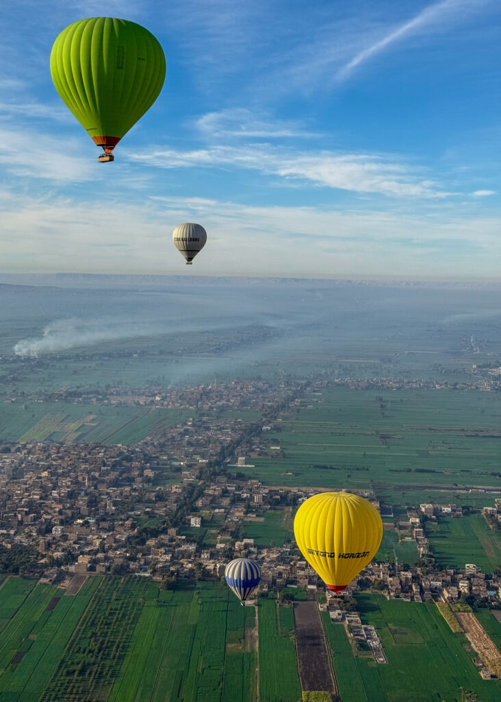 Hot air balloons floating over the lush green fields and villages of Luxor, Egypt, during sunrise.