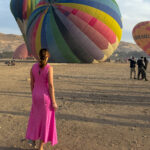 Woman in a pink dress standing in front of a colorful hot air balloon preparing for takeoff in Luxor, Egypt.