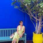 Woman in a pastel green dress sitting on a bench against the iconic blue wall of Le Jardin Majorelle in Marrakech, next to a vibrant yellow planter.