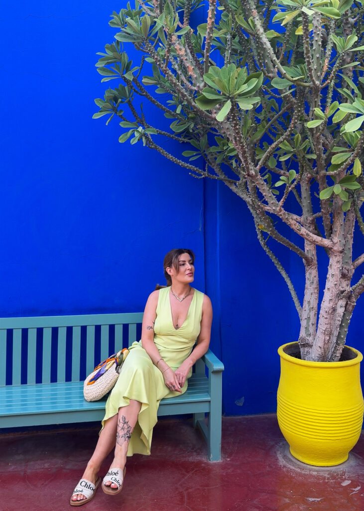 Woman in a pastel green dress sitting on a bench against the iconic blue wall of Le Jardin Majorelle in Marrakech, next to a vibrant yellow planter.
