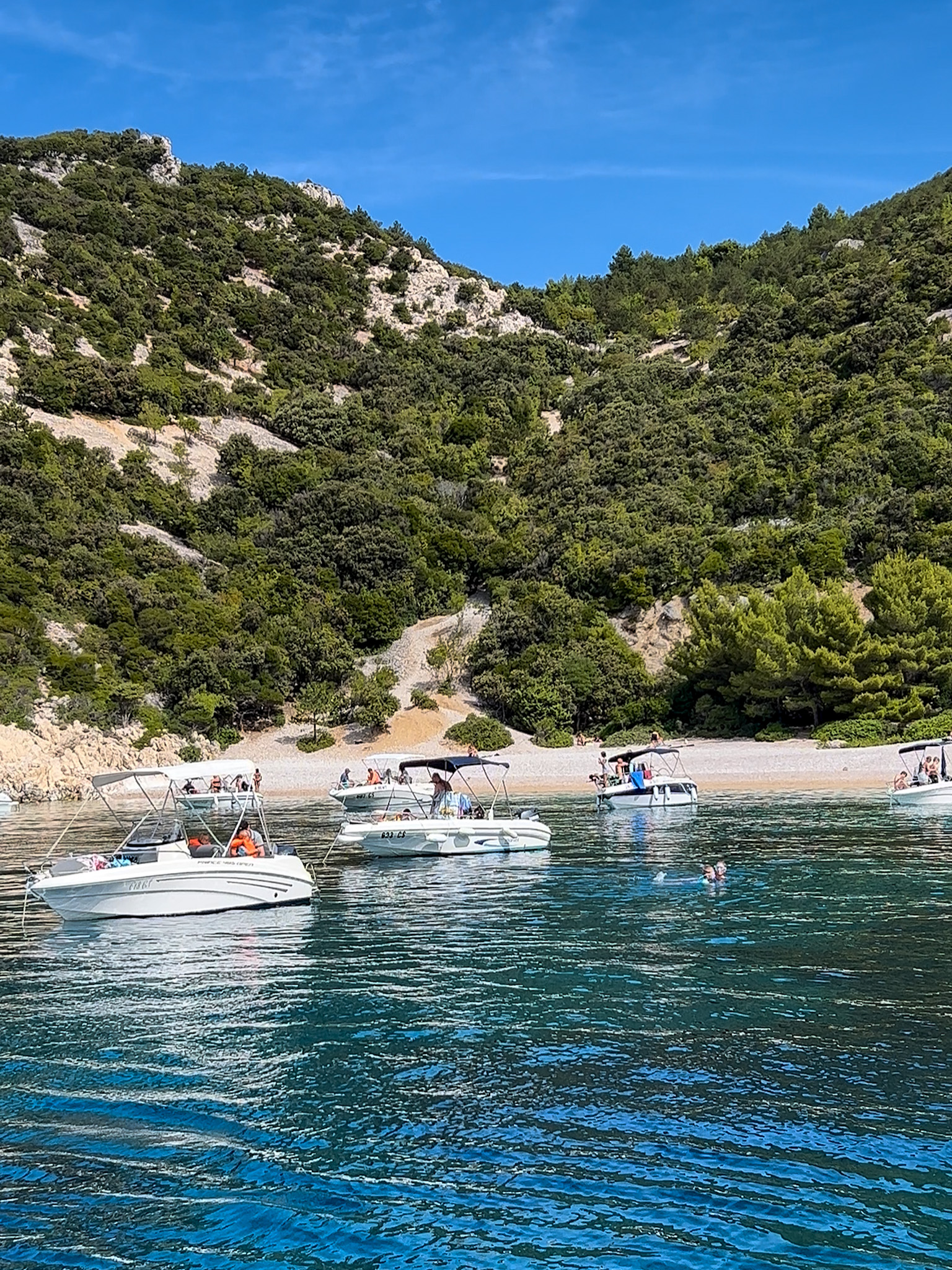 Lubenice Beach on Cres Island, Croatia, featuring turquoise waters, lush green hills, and boats anchored near the secluded pebbled shoreline.