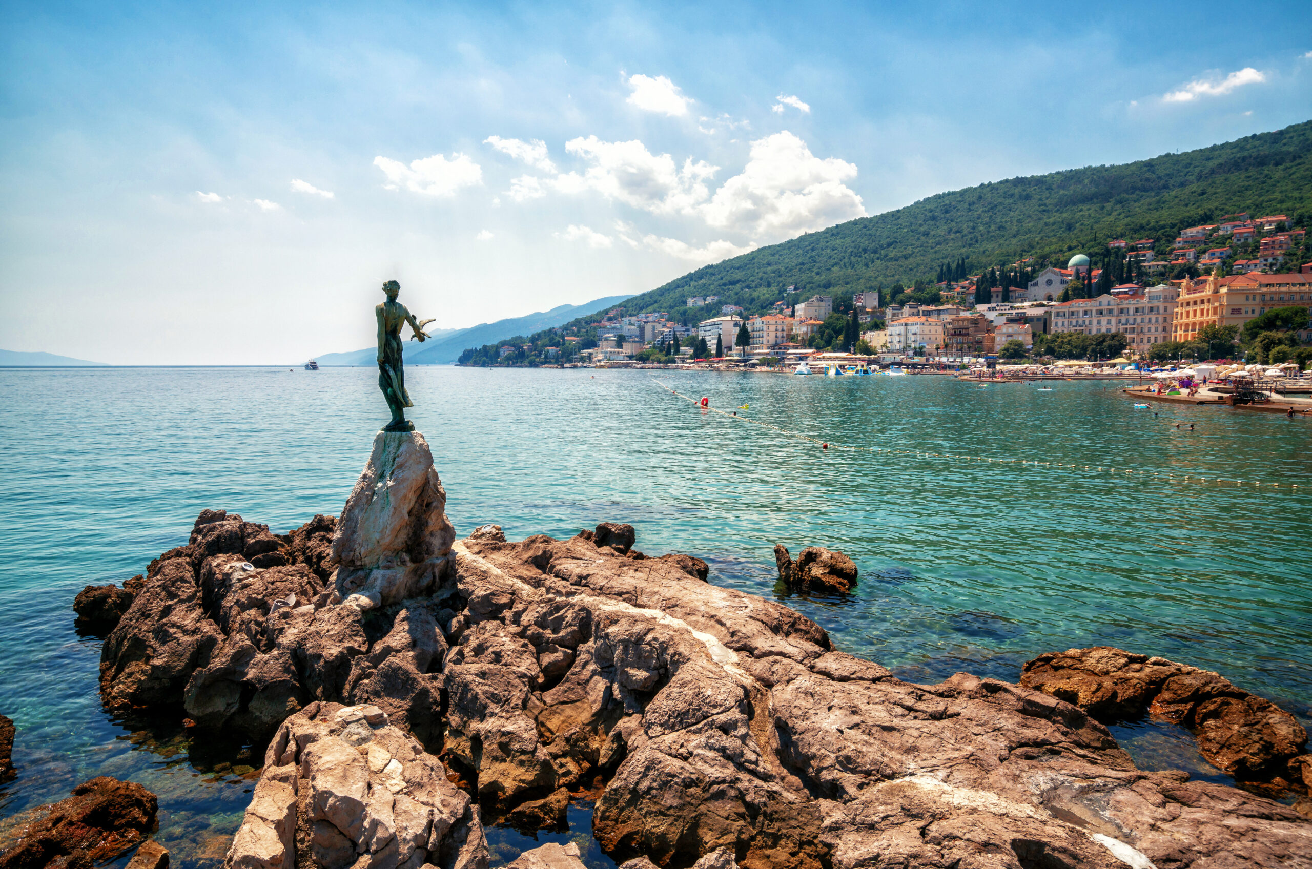 Statue of Maiden with the Seagull on the rocky Adriatic coast in Opatija, Croatia, with the town and lush green hills in the background.