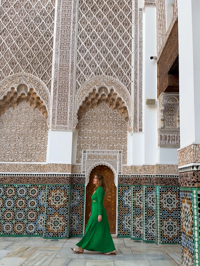 Woman in a green dress walking through the intricate tiled courtyard of Medersa Ben Youssef in Marrakech.