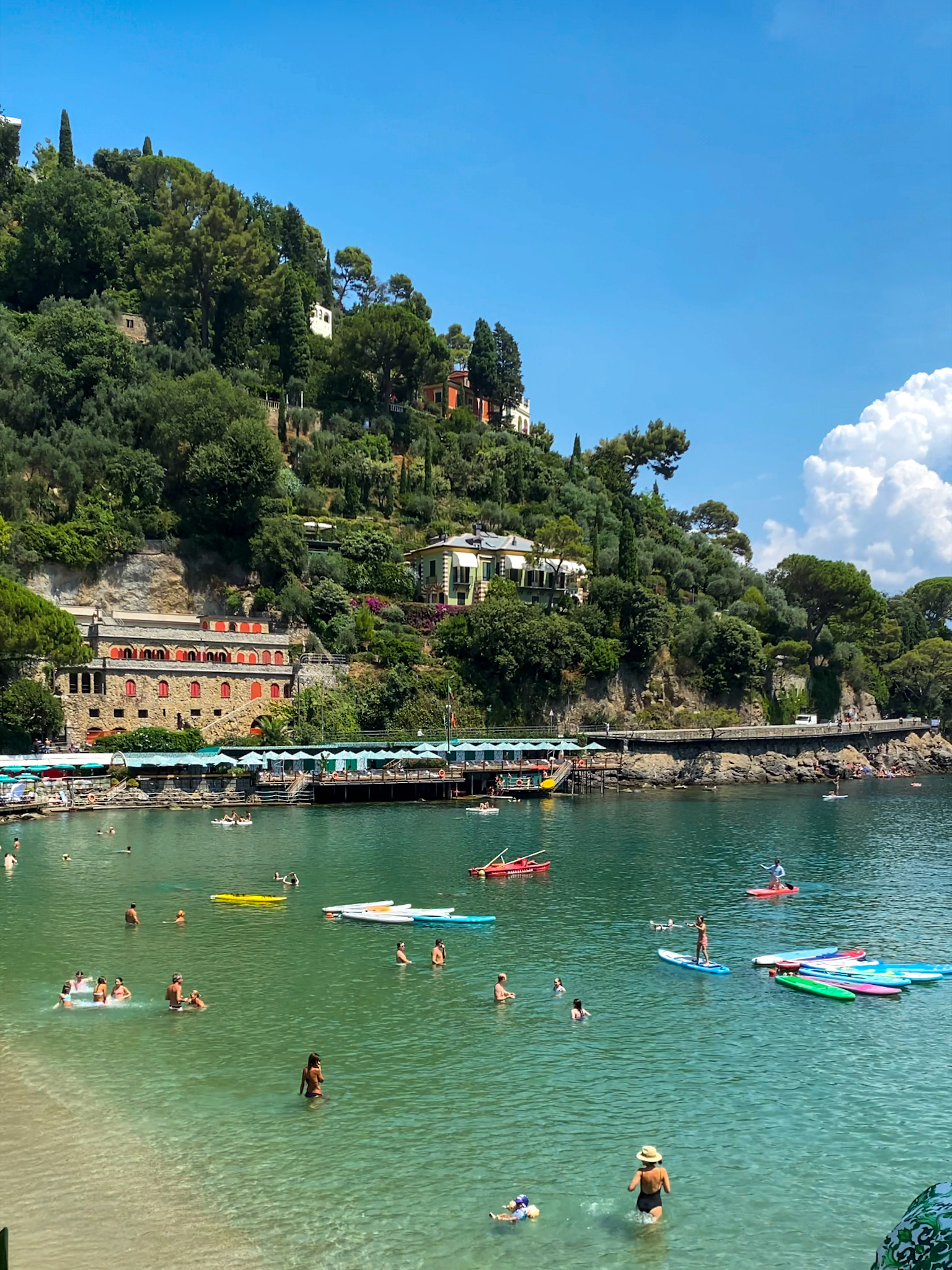 A lively beach scene at Paraggi Beach, with turquoise waters, people swimming, and paddleboards floating under the Italian sun.