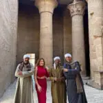 Philae Temple in Aswan, Egypt, featuring ancient Egyptian columns and carvings, with a traveler in a red dress posing with local Nubian men.