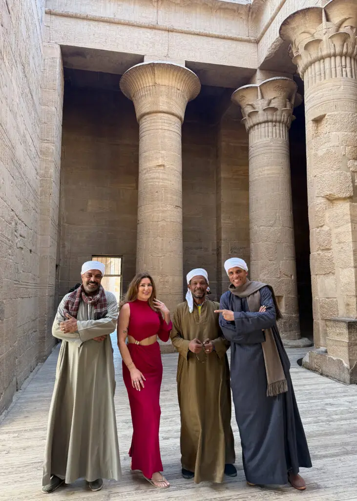 Philae Temple in Aswan, Egypt, featuring ancient Egyptian columns and carvings, with a traveler in a red dress posing with local Nubian men.