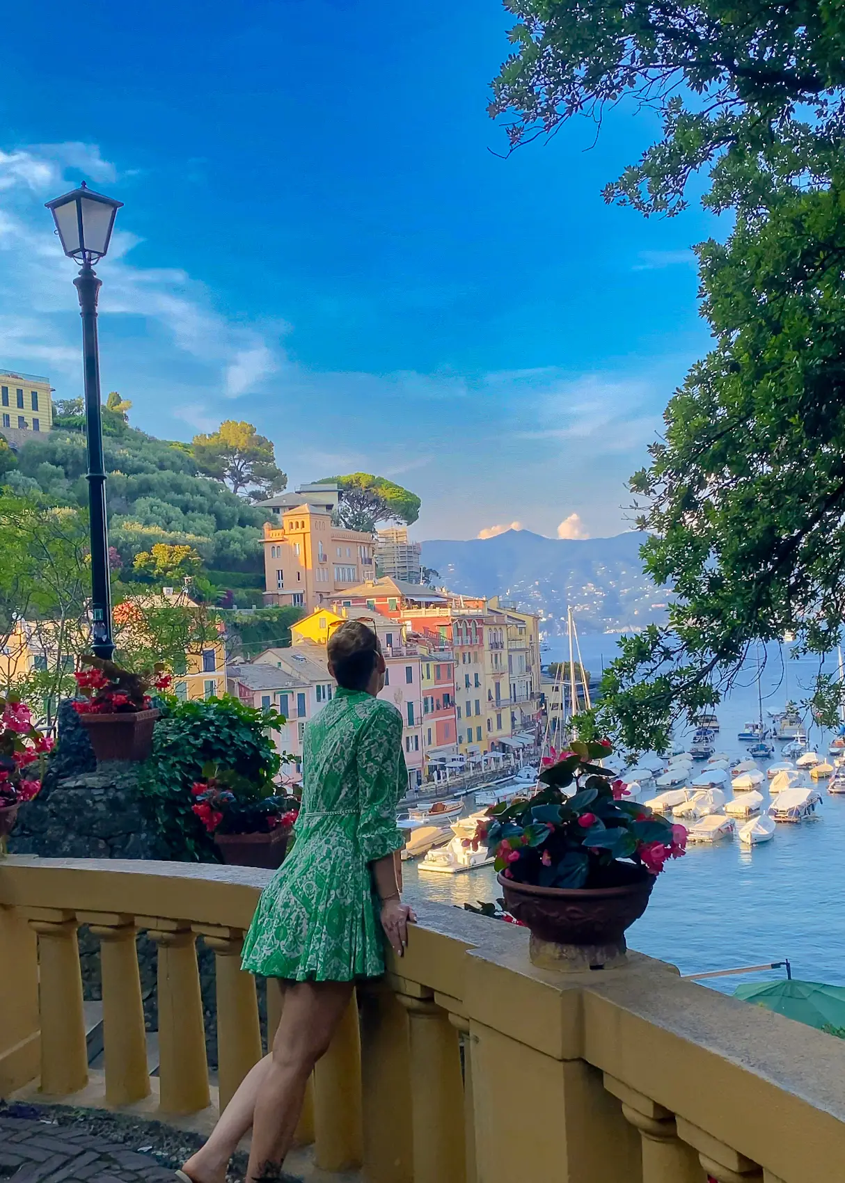 A woman in a green dress admiring the stunning Portofino harbor from a scenic viewpoint, with colorful buildings and yachts in the background.