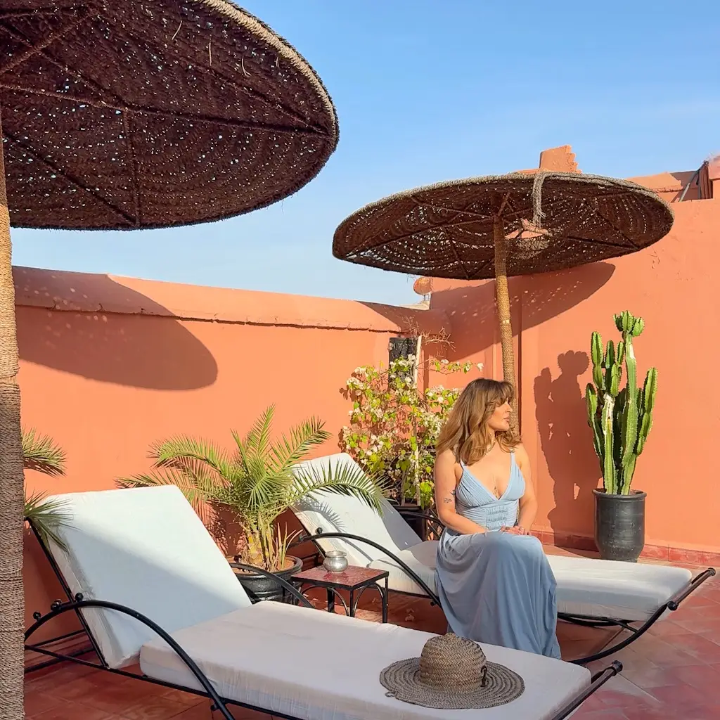 A woman in a light blue dress sits on a sun lounger at the rooftop terrace of Riad Dar Dialkoum in Marrakech, Morocco, surrounded by terracotta walls, lush greenery, and woven parasols under the warm Moroccan sun.