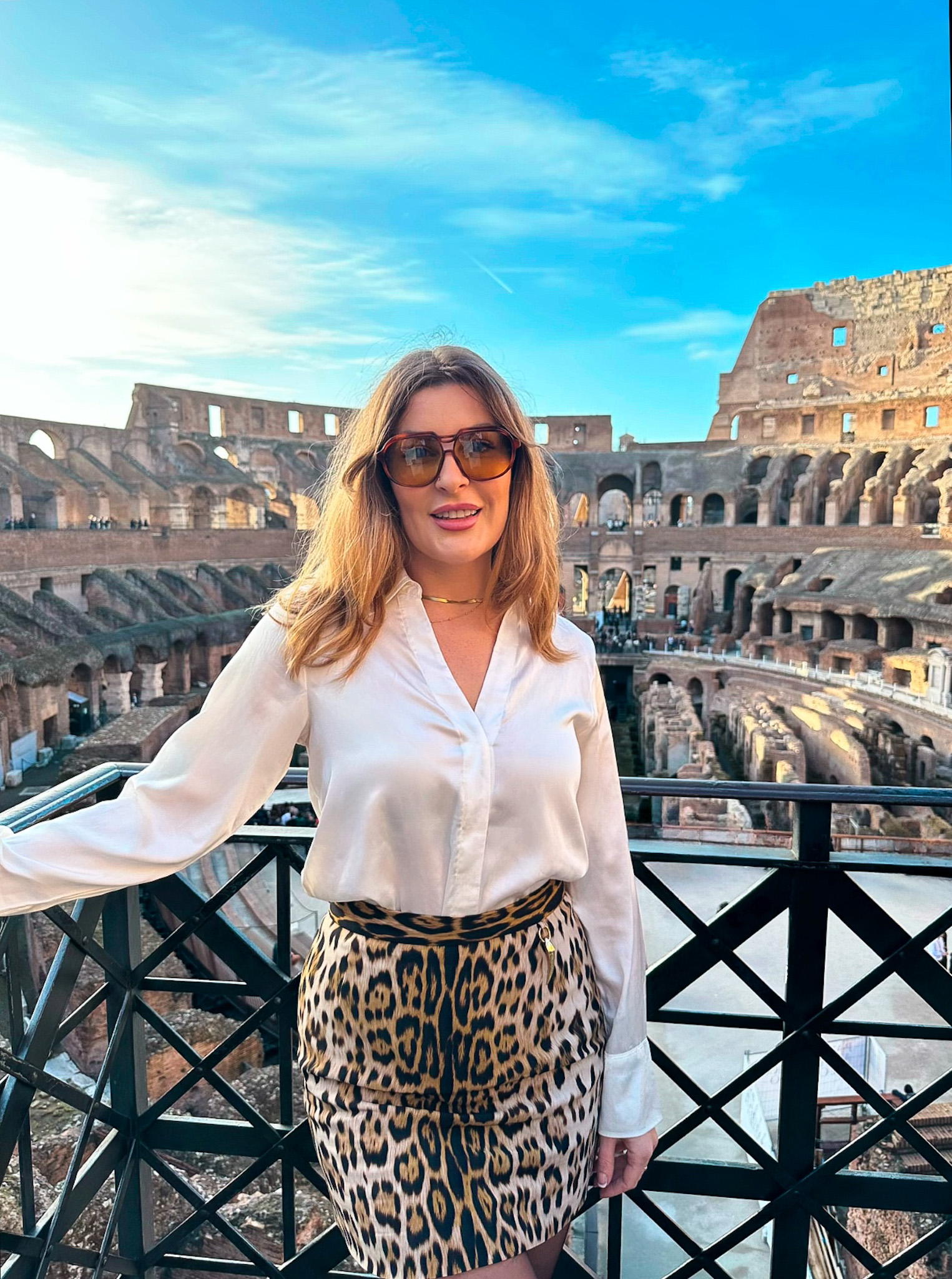 A woman in sunglasses, a white blouse, and a leopard-print skirt posing in front of the ancient Colosseum in Rome, with the historic amphitheater’s arches and stone structure visible in the background.