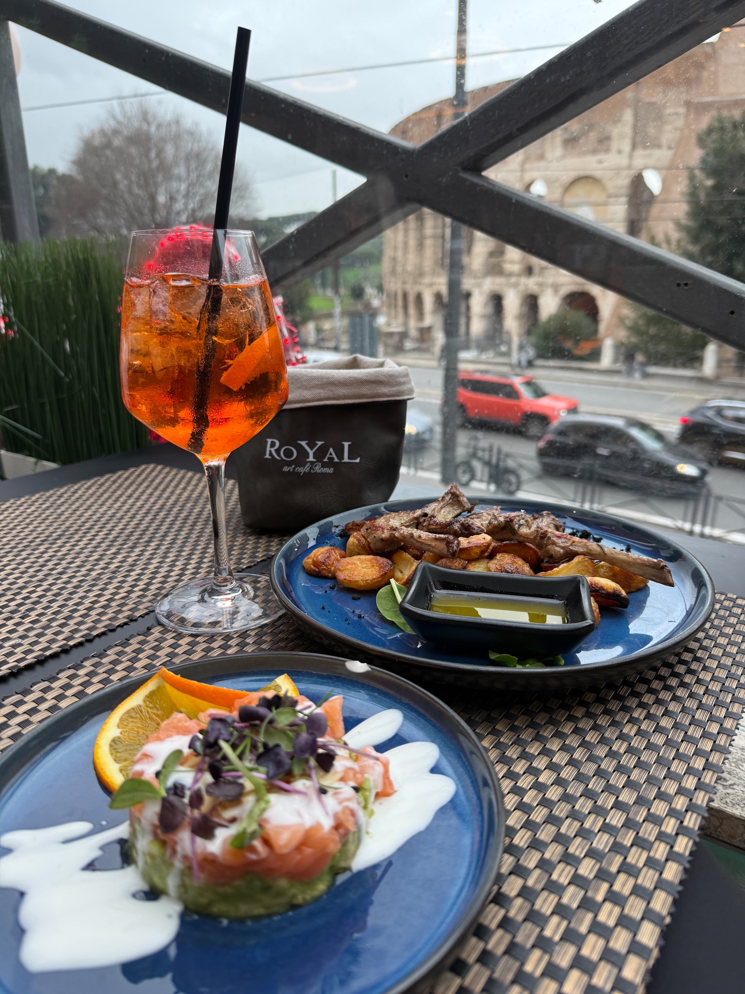 A table at Royal Art Caffè Roma featuring a vibrant Aperol Spritz, salmon tartare, and grilled lamb with roasted potatoes, overlooking the Colosseum in Rome.