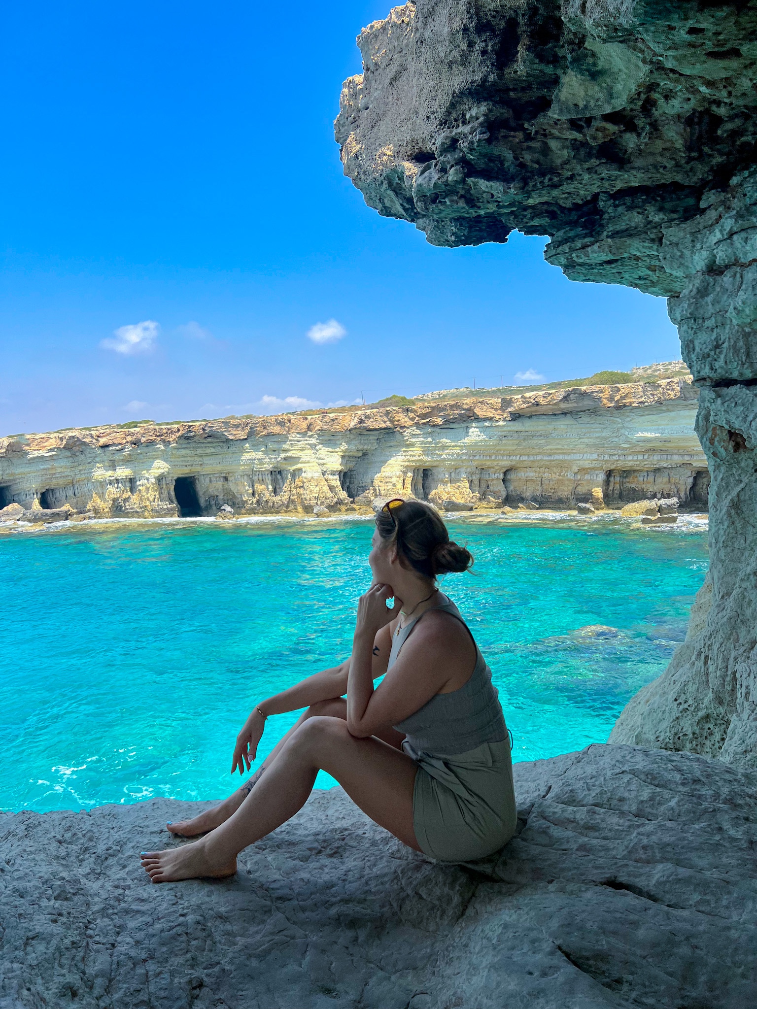 A woman sitting on the edge of a rock at Ayia Napa sea caves, gazing at the clear blue waters with the stunning limestone cliffs in the background.