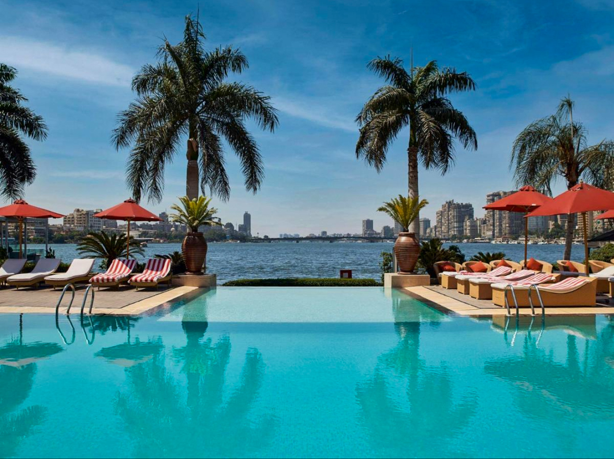 A luxurious infinity pool at Sofitel Cairo Nile, overlooking the river with palm trees, red sun umbrellas, and a stunning city skyline in the background.