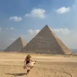 A woman walking with a flowing scarf in front of the Great Pyramids of Giza under a clear blue sky.