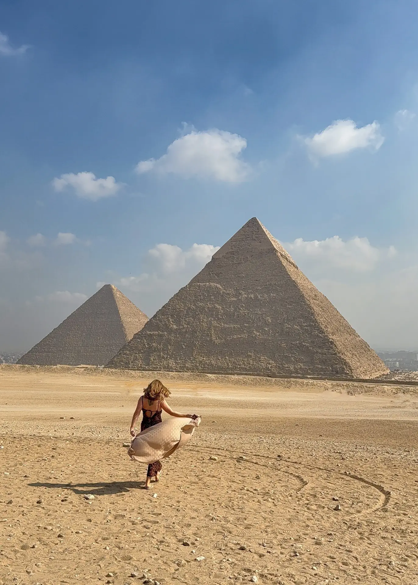 A woman walking with a flowing scarf in front of the Great Pyramids of Giza under a clear blue sky.