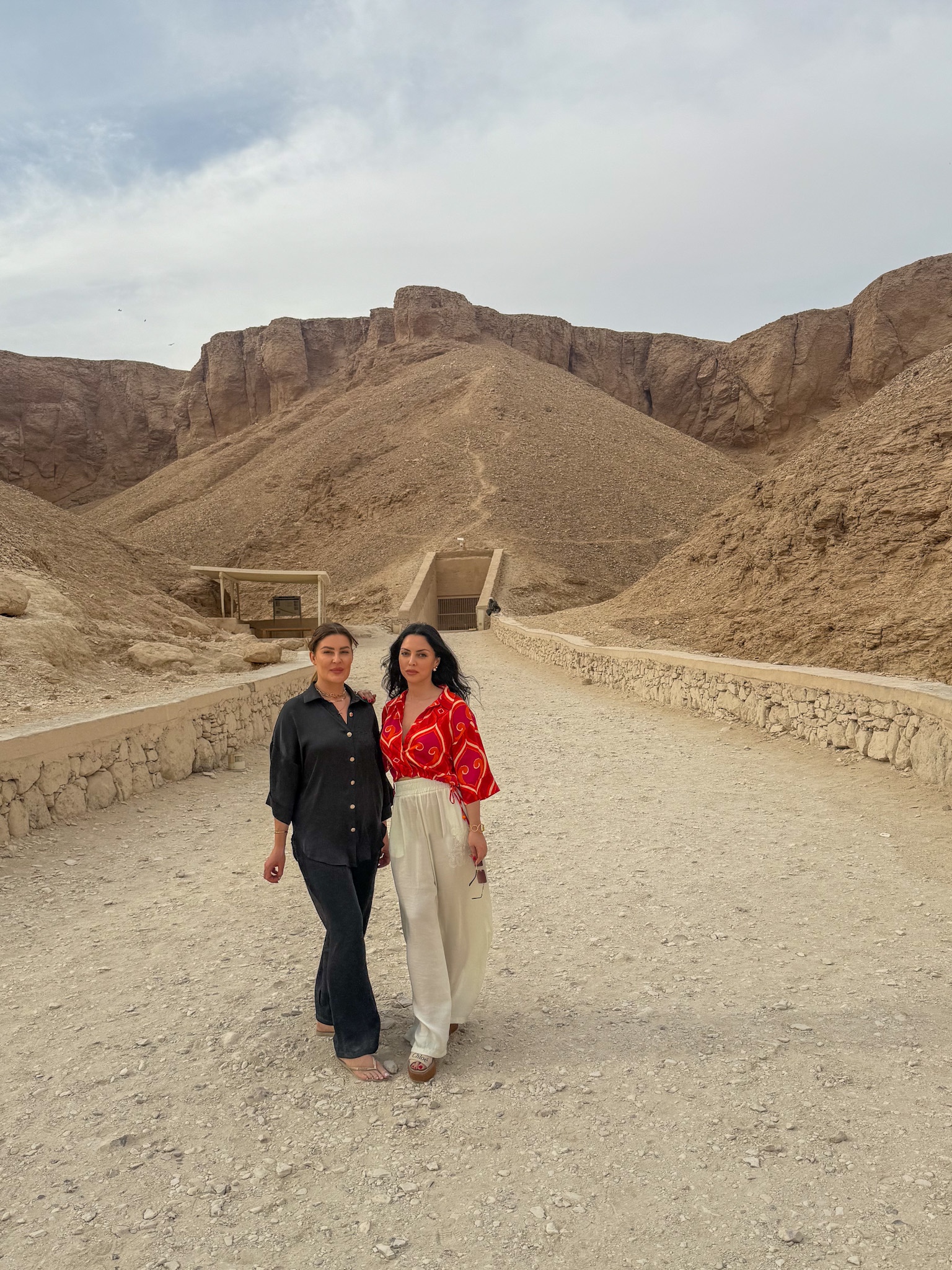 Two women walking towards the entrance of a tomb in the Valley of the Kings, Luxor, surrounded by rugged desert mountains.