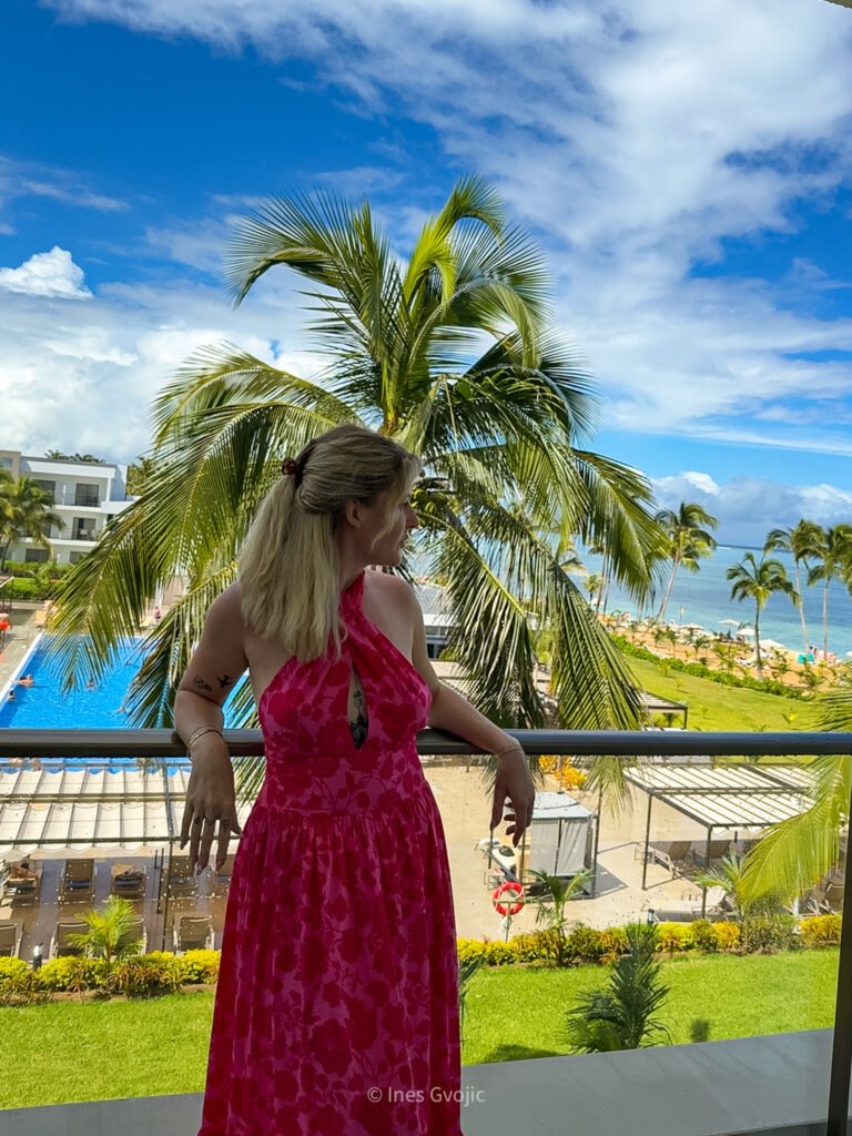 Woman in pink dress standing on balcony overlooking pool, palm trees, and beach at Riu Palace Hotel Mauritius.