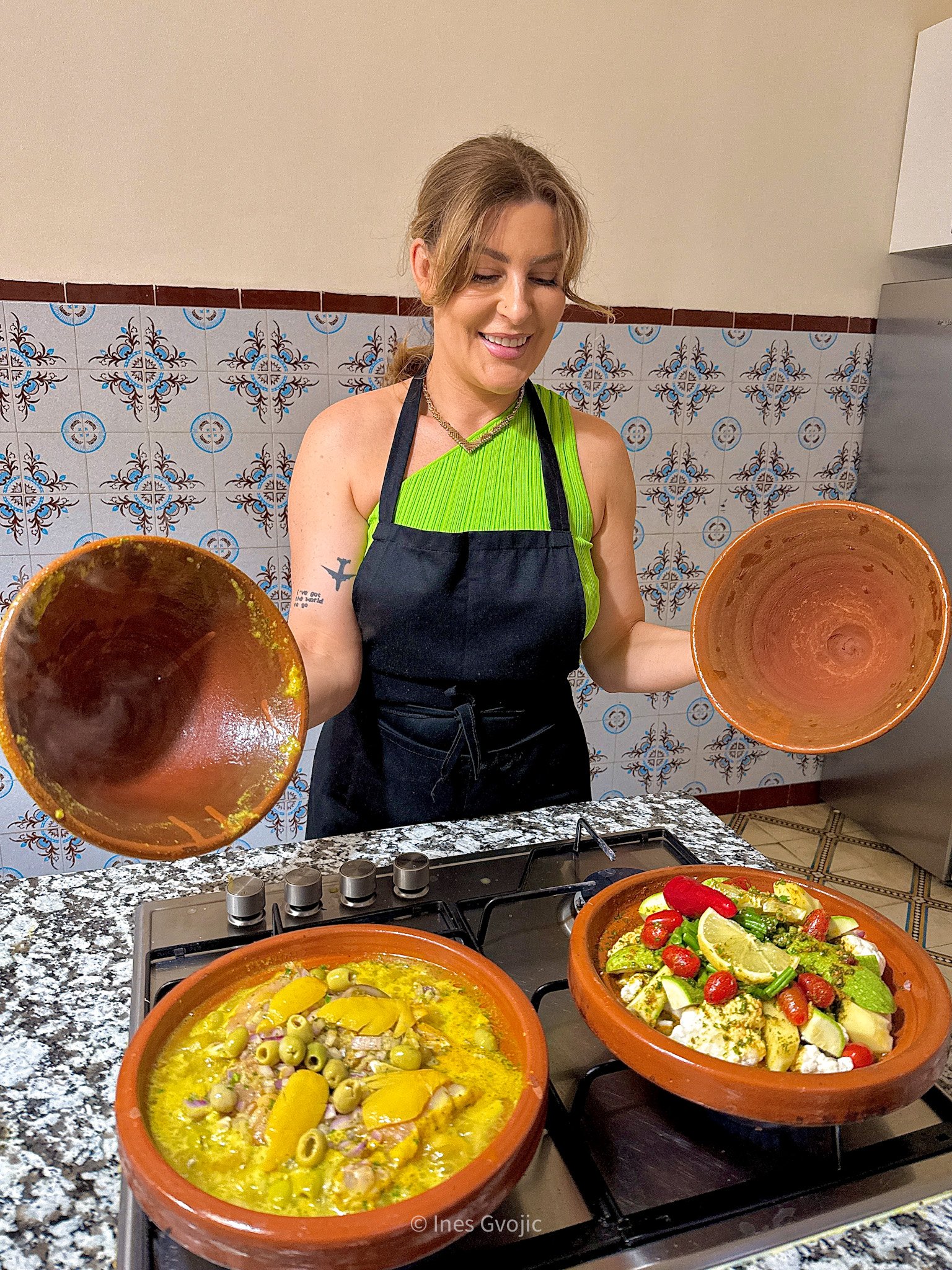 Woman in apron holding tagine lids, standing in a Moroccan kitchen with two clay pots filled with traditional tagine dishes.