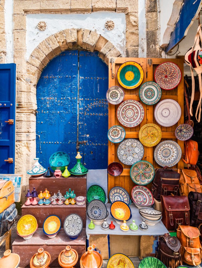Display of handmade Moroccan plates, ceramics, and leather bags set against a blue arched door in Marrakech.