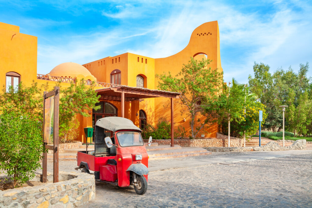 Red tuk-tuk parked in front of bright orange building in El Gouna, Egypt, surrounded by trees and desert landscaping.