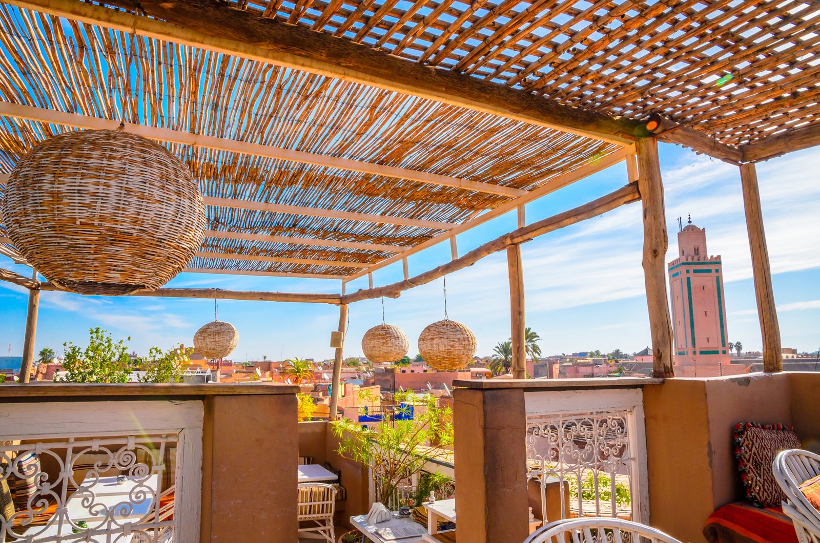 Traditional Marrakech rooftop terrace with woven lanterns, shaded seating, and city view with mosque tower.