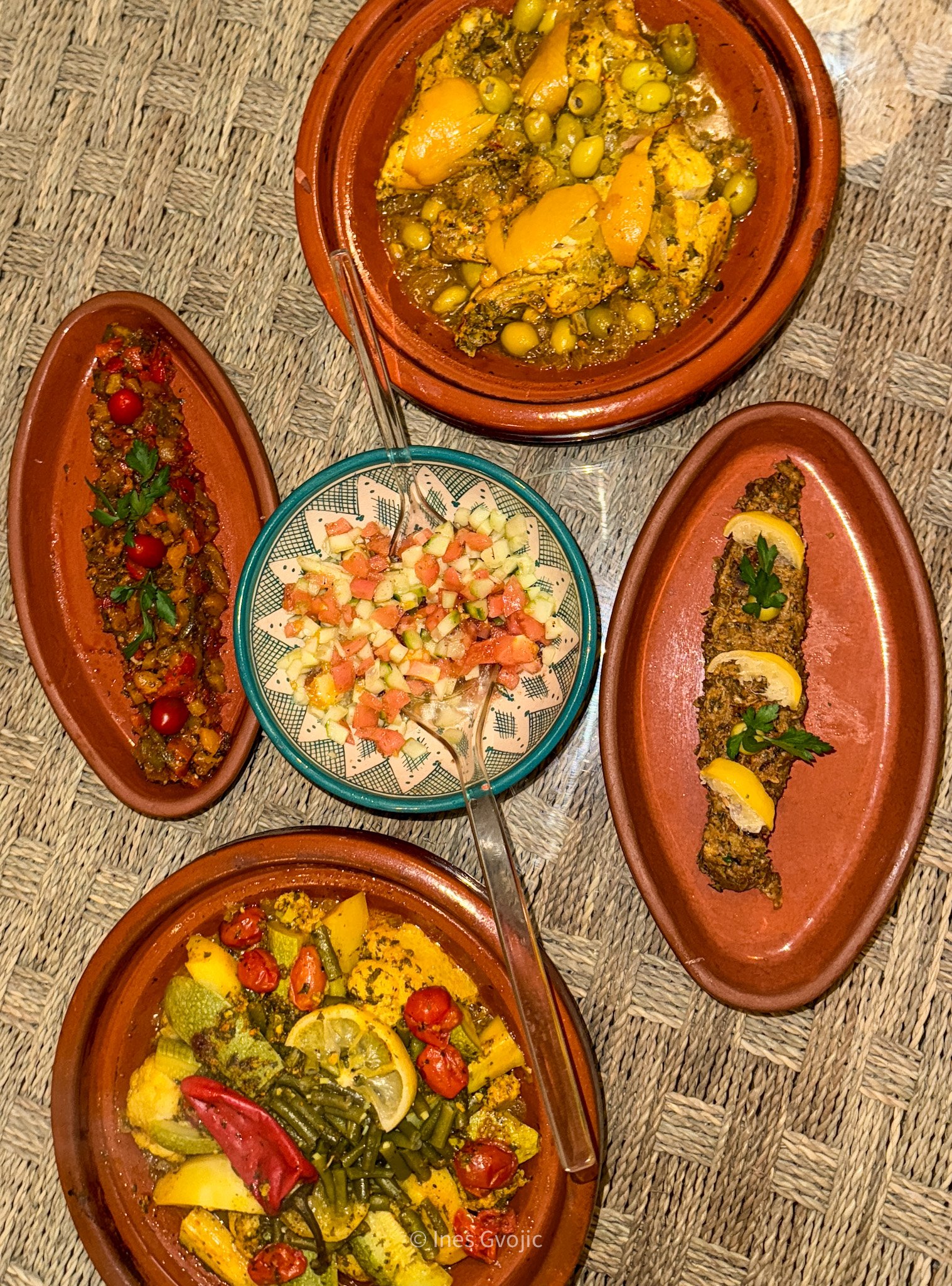 Top-down view of assorted Moroccan tagines with chicken, vegetables, and fish, served with salads on a woven table surface.