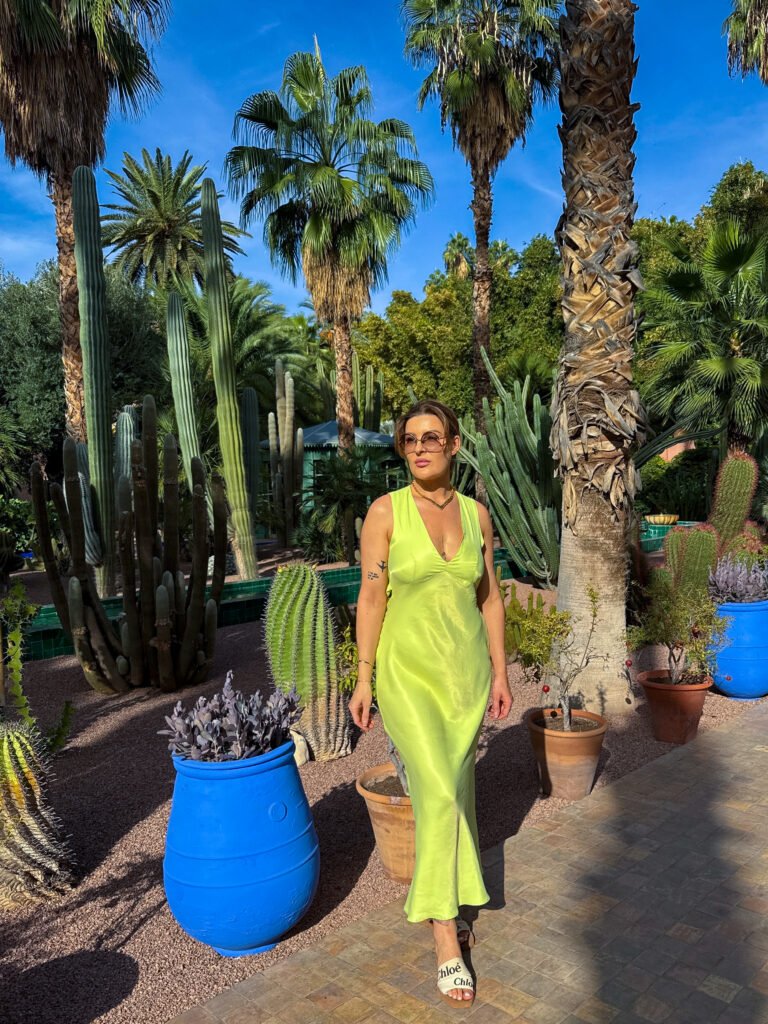 Woman in a lime-green dress walking through Jardin Majorelle in Marrakech, surrounded by cactuses, palm trees, and iconic blue pots.