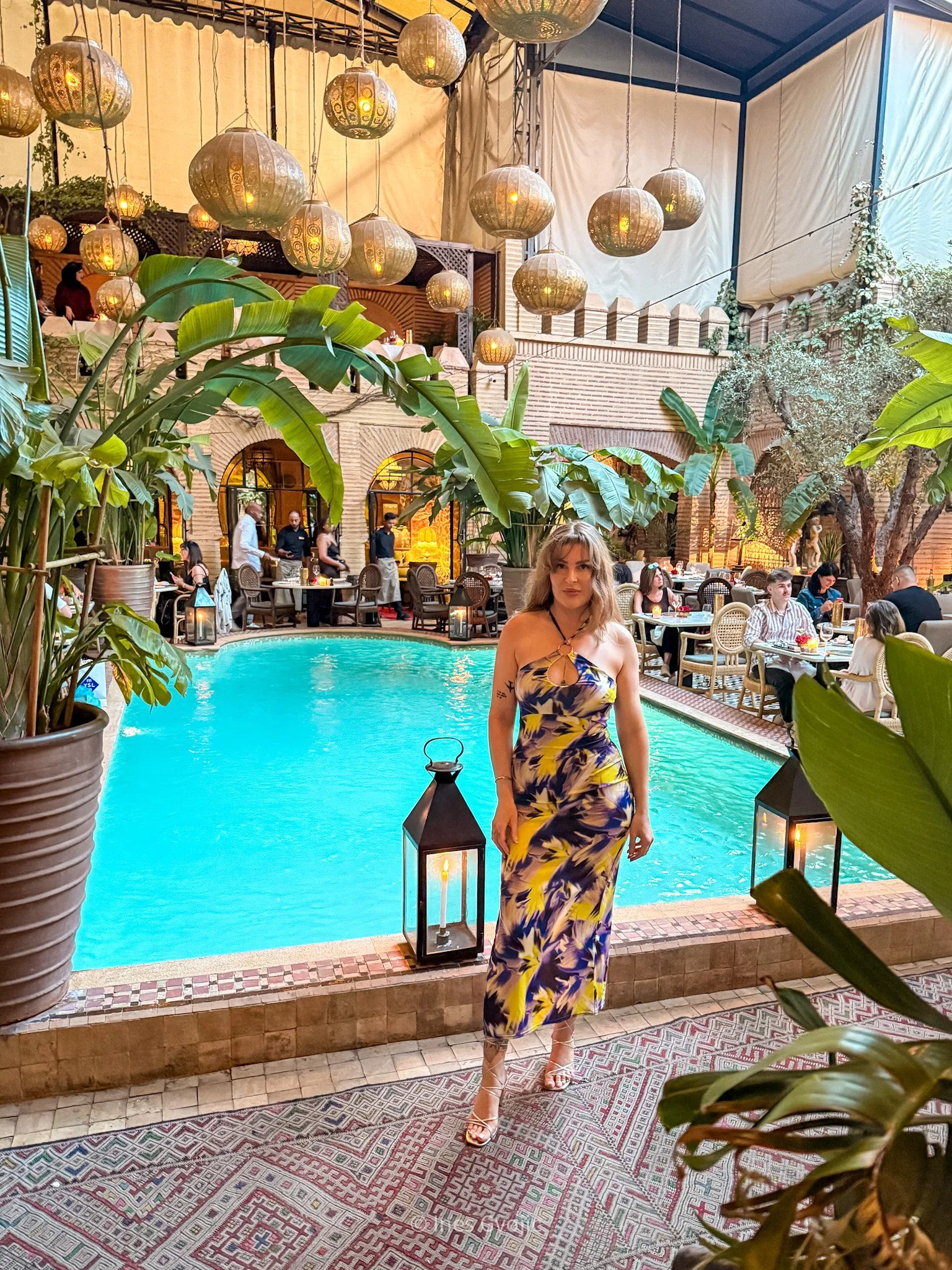 Woman in a printed dress standing beside the pool at La Trattoria Marrakech, surrounded by tropical plants and lanterns.