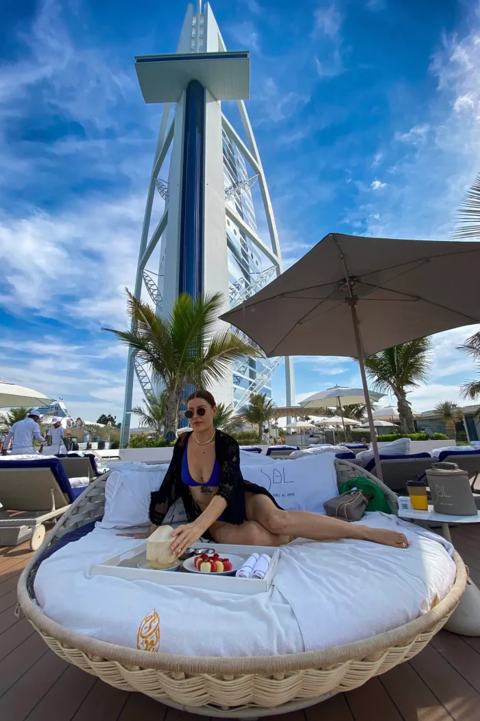 A woman relaxing on a sunbed at a luxury beach club with the iconic Burj Al Arab in Dubai towering in the background.